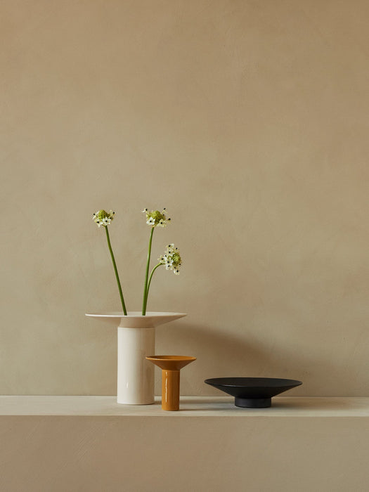 A tall off-white Hana Vase with three flower stems, next to a small yellow vase and a low black bowl, on a shelf.