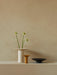 A tall off-white Hana Vase with three flower stems, next to a small yellow vase and a low black bowl, on a shelf.