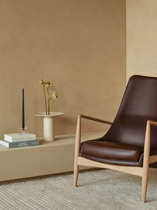Side view of The Seal High Back chair in brown leather. Nearby, a cream Hana Vase with flowers sits on a ledge with books.