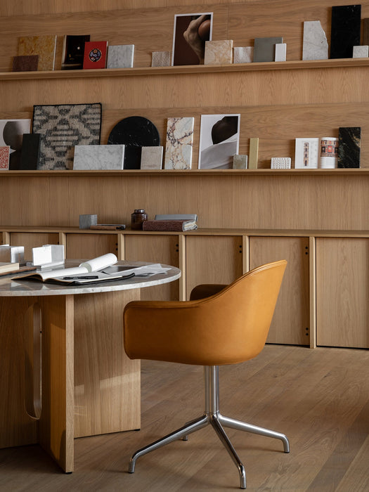 A tan Harbour Dining Chair at a marble-topped Androgyne Dining Table in a studio with wooden shelves of material samples.