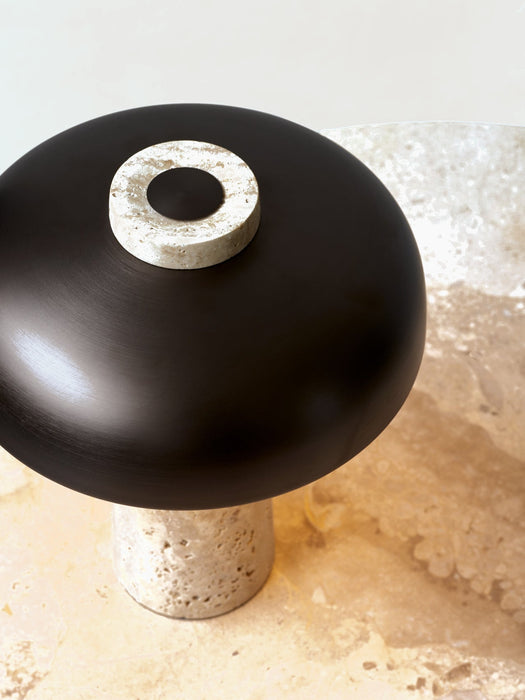 A close-up of the Reverse Table Lamp with a travertine stone base and black shade, resting on a stone Harbour Column Table.