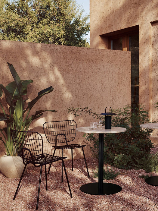 A Harbour Column Dining Table and two black WM String Dining Chairs on a sunlit gravel patio with a Ray Table Lamp.