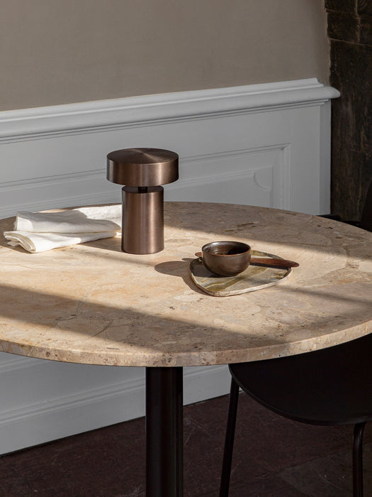 A Harbour Column Dining Table with a round Kunis Breccia top is bathed in sunlight, styled with a bronze lamp and teacup.