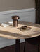 A Harbour Column Dining Table with a round Kunis Breccia top is bathed in sunlight, styled with a bronze lamp and teacup.