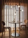 A round Harbour Column Dining Table set with two glasses and a carafe, placed between two wooden chairs under a chandelier.