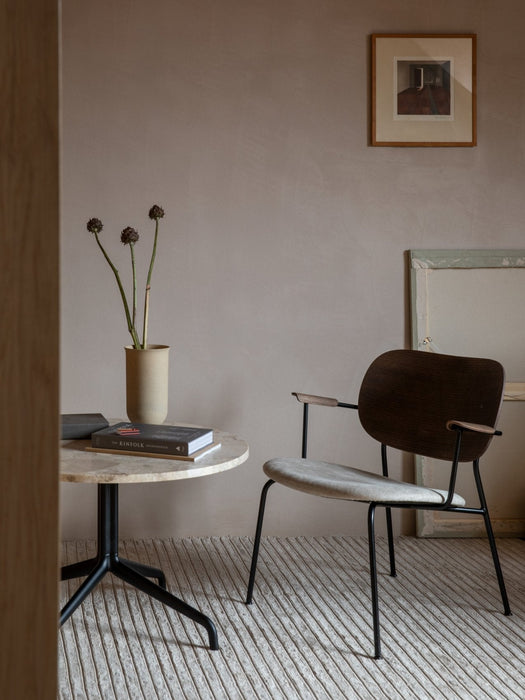 A Co Lounge Chair beside a Harbour Column Lounge Table with a Cyclades Vase and books on a Gravel Rug in a softly lit room.