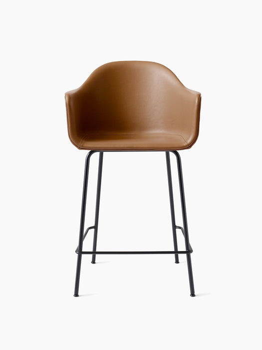 A Harbour Counter Chair seen from the front, with a tan leather upholstered shell, armrests, and a tall black steel base.
