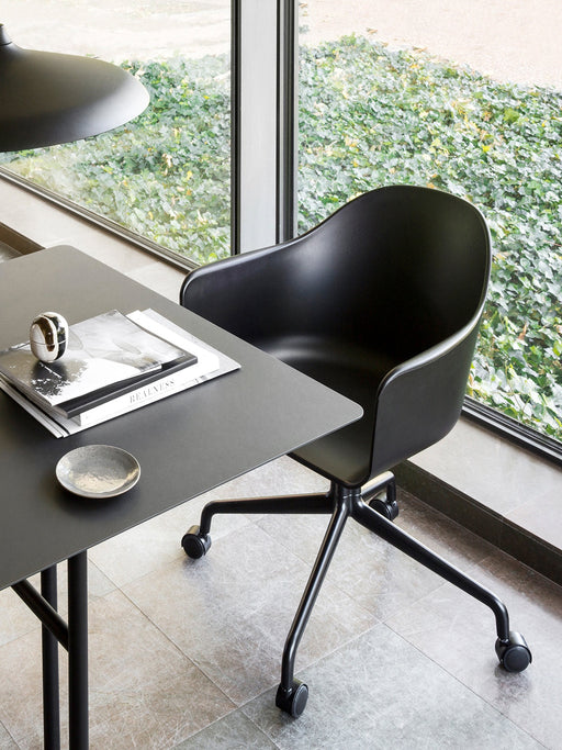 The Harbour Dining Chair, with a black star base and casters, is shown at a black desk next to a window with a garden view.