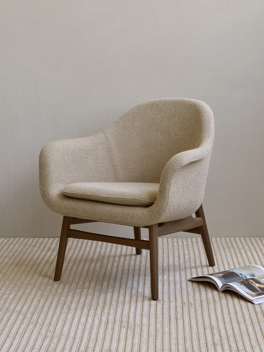 A beige Harbour Lounge Chair with a dark wood frame on a striped rug in a minimalist room, with a magazine on the floor.
