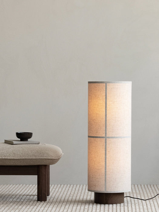 A lit Hashira Floor Lamp stands on a ribbed Houkime Rug next to a Brasilia Ottoman holding a book and a small NNDW Cup.