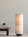 A lit Hashira Floor Lamp stands on a ribbed Houkime Rug next to a Brasilia Ottoman holding a book and a small NNDW Cup.
