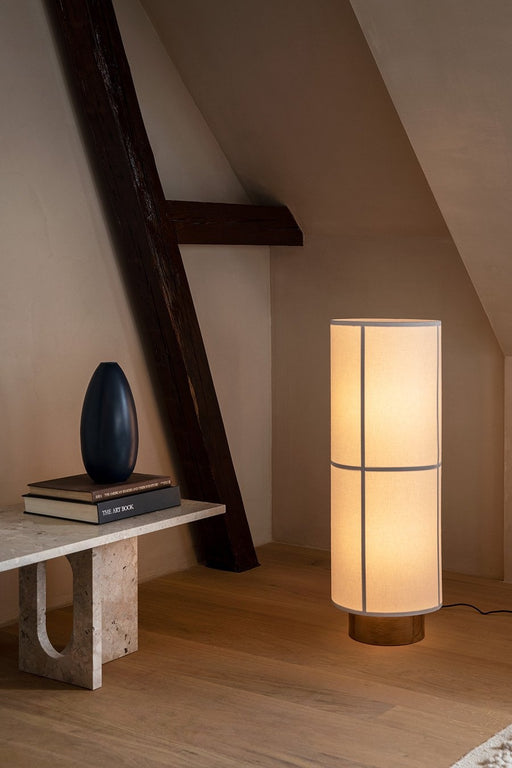 A lit Hashira Floor Lamp stands on a wooden floor in a cosy attic room, next to a low stone table with books and a dark vase.