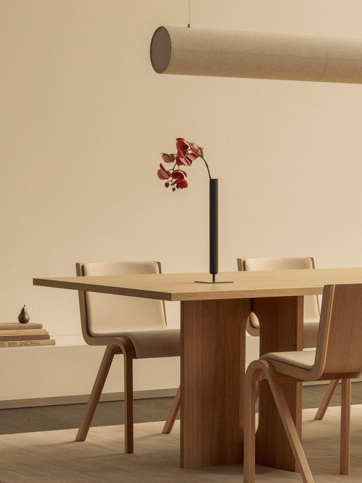 The beige Hashira Linear Pendant hangs above a wooden Androgyne Dining Table set with Ready Dining Chairs and a Stance Vase.