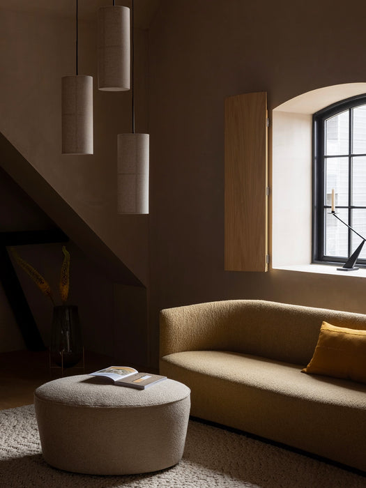 Three Hashira Pendant Lamps hang in a dimly lit room over a yellow Tearoom Sofa and a Cairn Pouf next to an arched window.