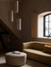 Three Hashira Pendant Lamps hang in a dimly lit room over a yellow Tearoom Sofa and a Cairn Pouf next to an arched window.