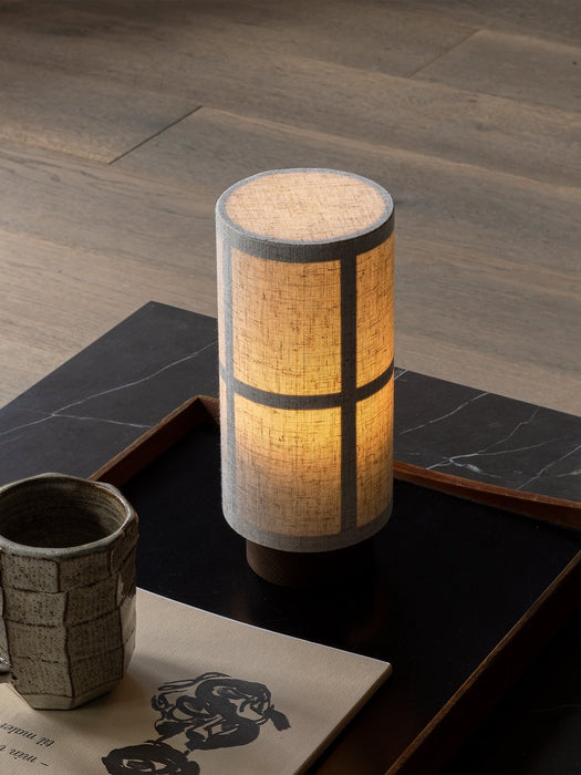The glowing Hashira Table Lamp, Portable on a dark wooden tray next to a ceramic mug and book on a black marble plinth.