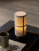 The glowing Hashira Table Lamp, Portable on a dark wooden tray next to a ceramic mug and book on a black marble plinth.