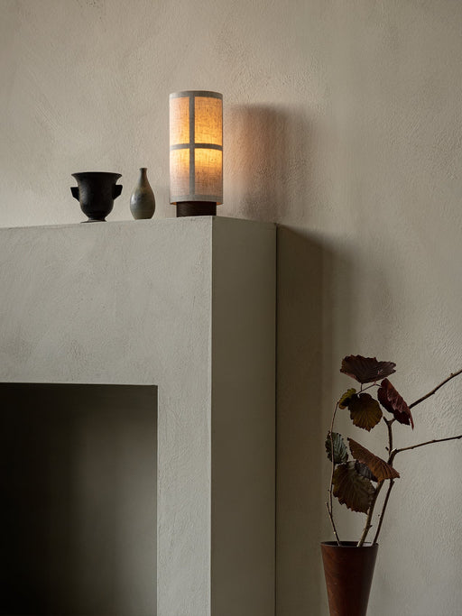 The Hashira Table Lamp, Portable, glows warmly on a grey mantelpiece next to two small vases, against a textured beige wall.