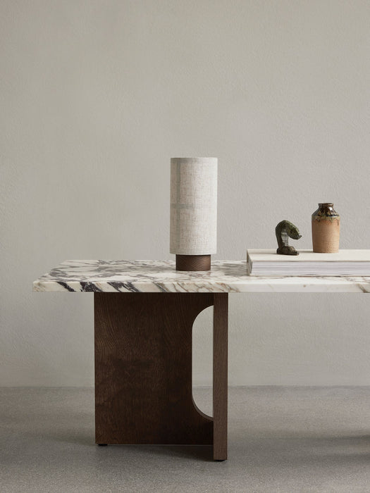 Hashira Portable Table Lamp with a linen shade sits on a marble Androgyne Lounge Table next to a book and decorative items.