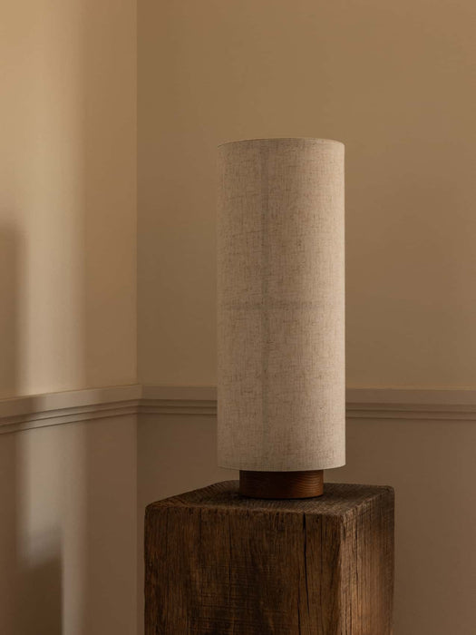 The Hashira Table Lamp, Raw, with its cylindrical linen shade and ash base, sitting on a rustic wooden block in a room corner