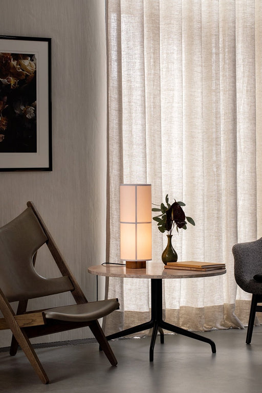 The Hashira Table Lamp emits a warm glow on a marble side table, placed next to a wooden Knitting Chair in a modern interior.