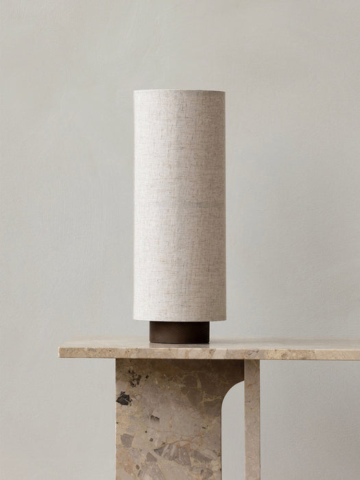 The Hashira Table Lamp, Raw, shown with its cylindrical linen shade and dark wood base, sits on a beige marble console table.