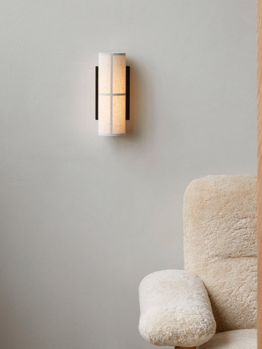 The Hashira Wall Lamp in raw linen, glowing on a light grey wall next to the fluffy, cream-coloured Brasilia Lounge Chair.