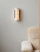 The Hashira Wall Lamp in raw linen, glowing on a light grey wall next to the fluffy, cream-coloured Brasilia Lounge Chair.