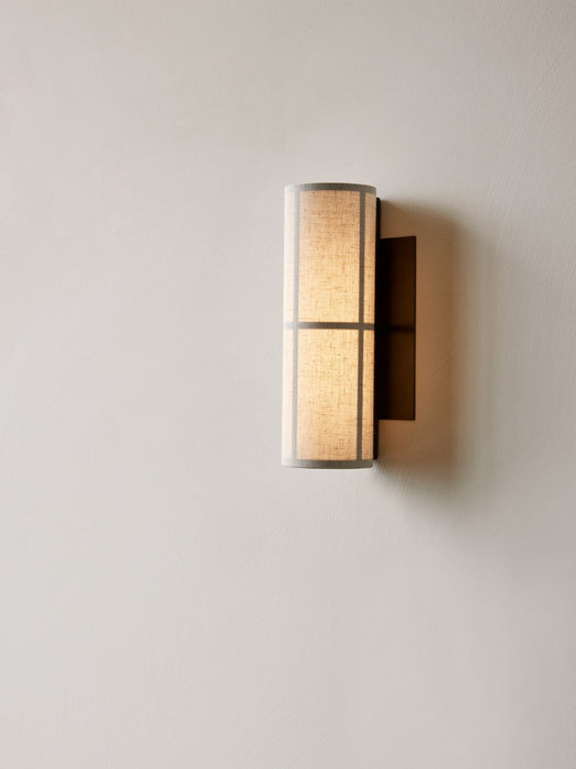 A close-up of the Hashira Wall Lamp with a raw linen shade, mounted on a light-coloured textured wall and emitting warm glow.