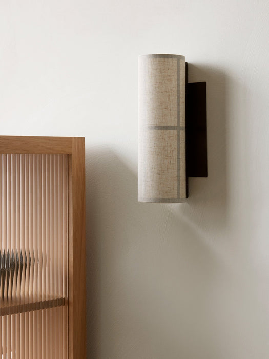 A close-up of the Hashira Wall Lamp in raw linen, mounted on a textured white wall next to a light oak fluted cabinet.