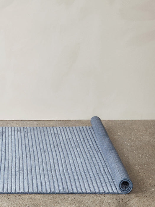 A blue ribbed Houkime Rug partially unrolled across a grey concrete floor, set against a plain off-white wall.