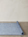 A blue ribbed Houkime Rug partially unrolled across a grey concrete floor, set against a plain off-white wall.
