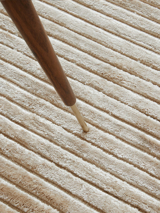 A detailed close-up of the beige Houkime Rug, highlighting its soft, linear texture under the tapered wooden leg of a chair.