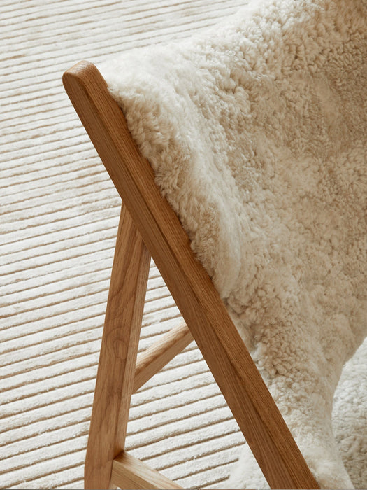 A detailed close-up of a wooden chair frame with a fluffy sheepskin throw, set against the ribbed texture of the Houkime Rug.