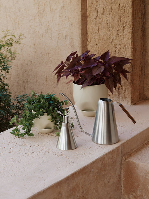 Stainless steel Hydrous Watering Can and Hydrous Mister sit on a stone ledge beside two plants in off-white Hydrous Planters.