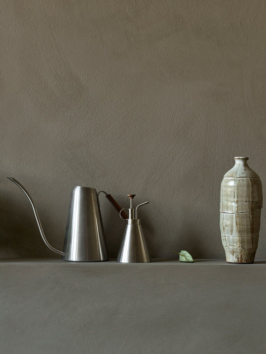 A sleek, brushed steel Hydrous Watering Can and Hydrous Plant Mister next to a rustic ceramic vase on a grey textured shelf.