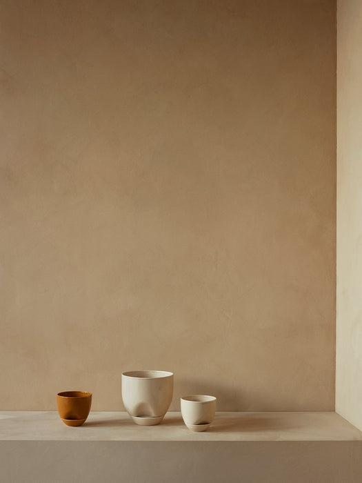 A trio of Hydrous Planters in varying sizes. One medium ochre planter and two off-white planters on a beige stone shelf.