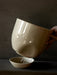 A hand holds the top of the glossy, off-white Hydrous Planter, showing the self-watering wick dangling into the water bowl.