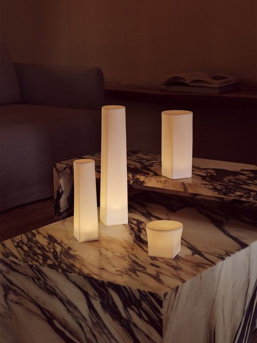 Four glowing Ignus Flameless Candles in various sculptural shapes, styled on tiered marble plinth tables in a dimly lit room.