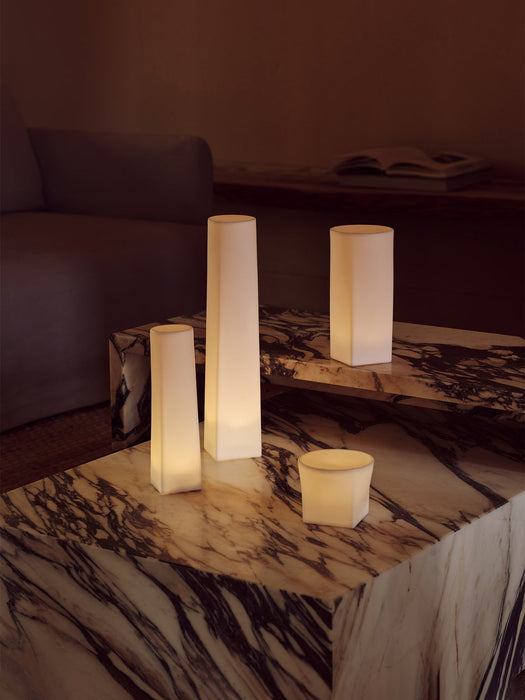 Four glowing Ignus Flameless Candles in various sculptural shapes, styled on tiered marble plinth tables in a dimly lit room.