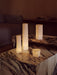 Four glowing Ignus Flameless Candles in various sculptural shapes, styled on tiered marble plinth tables in a dimly lit room.