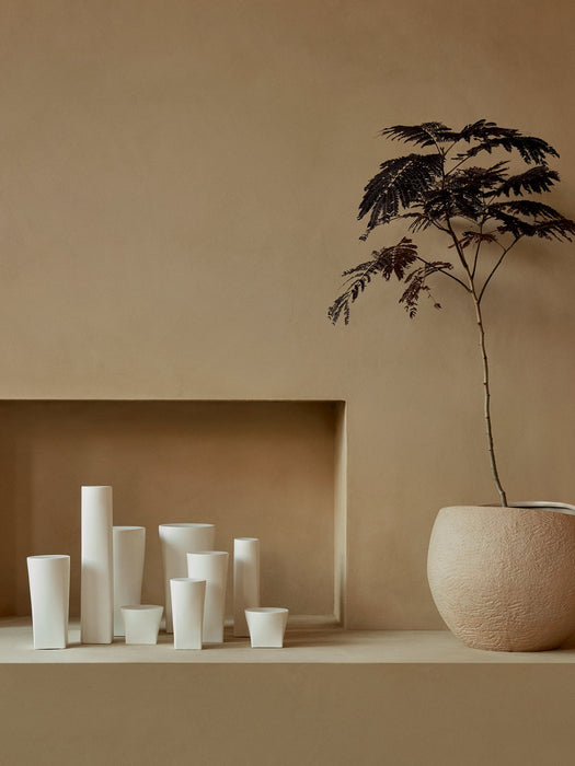 A collection of white Ignus Flameless Candles sits in a beige alcove next to a Plantas Planter holding a small tree.