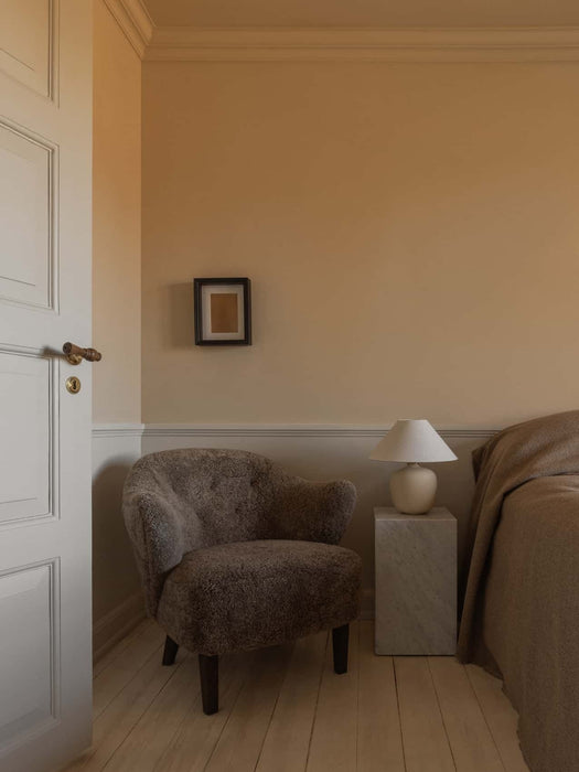 A cozy bedroom with a brown sheepskin Ingeborg Lounge Chair, a marble Plinth Tall side table and a cream Torso Table Lamp.