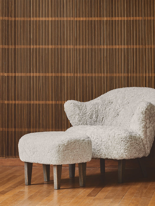 A white sheepskin Ingeborg Lounge Chair and footstool with wooden legs on a parquet floor in front of a wooden slatted wall.