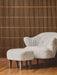 A white sheepskin Ingeborg Lounge Chair and footstool with wooden legs on a parquet floor in front of a wooden slatted wall.