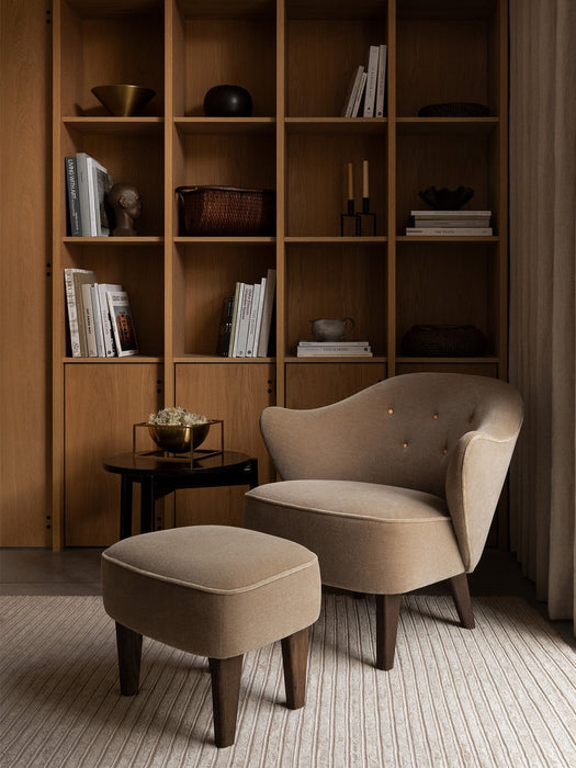 The Ingeborg Lounge Chair and Footstool in beige, beside a side table with a Kubus Bowl, in front of a wooden bookcase.