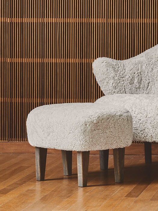 Close-up: Ingeborg Ottoman and part of the Lounge Chair, both in light sheepskin with dark oak legs on a hardwood floor.