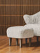 Close-up: Ingeborg Ottoman and part of the Lounge Chair, both in light sheepskin with dark oak legs on a hardwood floor.