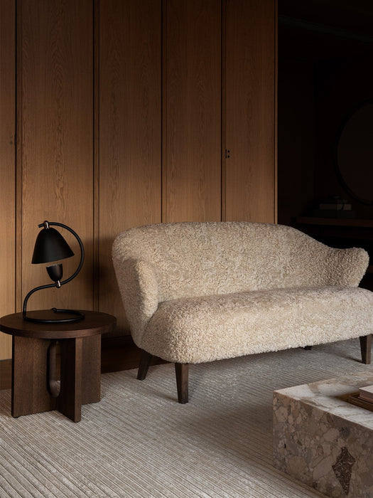 The cream sheepskin Ingeborg Sofa sits in a room with wood panelling, beside a dark wood table with a black Klampenborg Lamp.