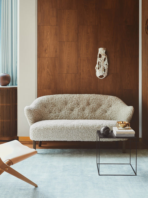 The white sheepskin Ingeborg Sofa on a light blue rug against a wood panelled wall, with a black Twin Table and a Saxe Chair.